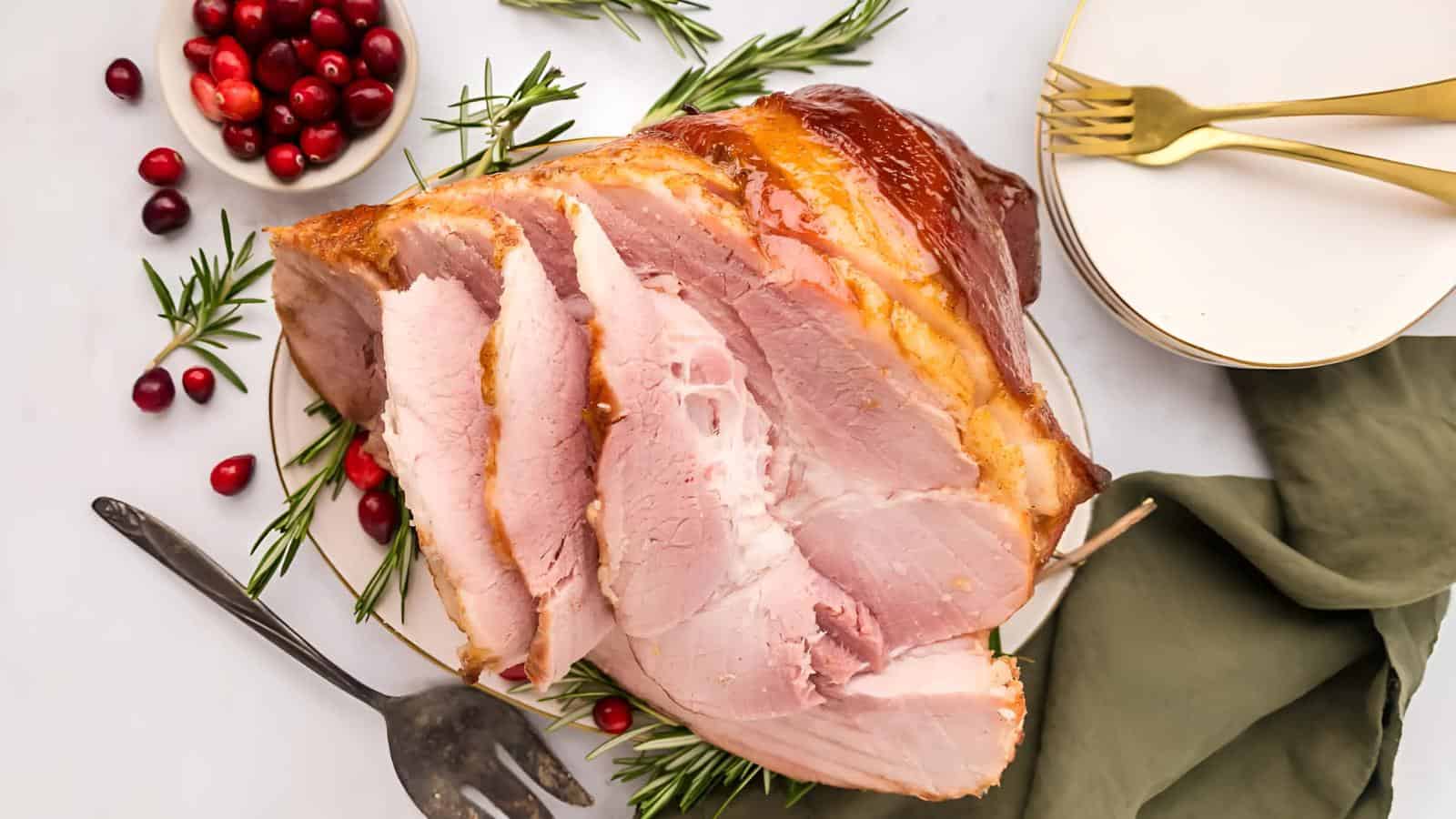 Sliced ham garnished with rosemary on a plate, near a bowl of cranberries, a serving fork, and stacked plates with gold forks on a green napkin.