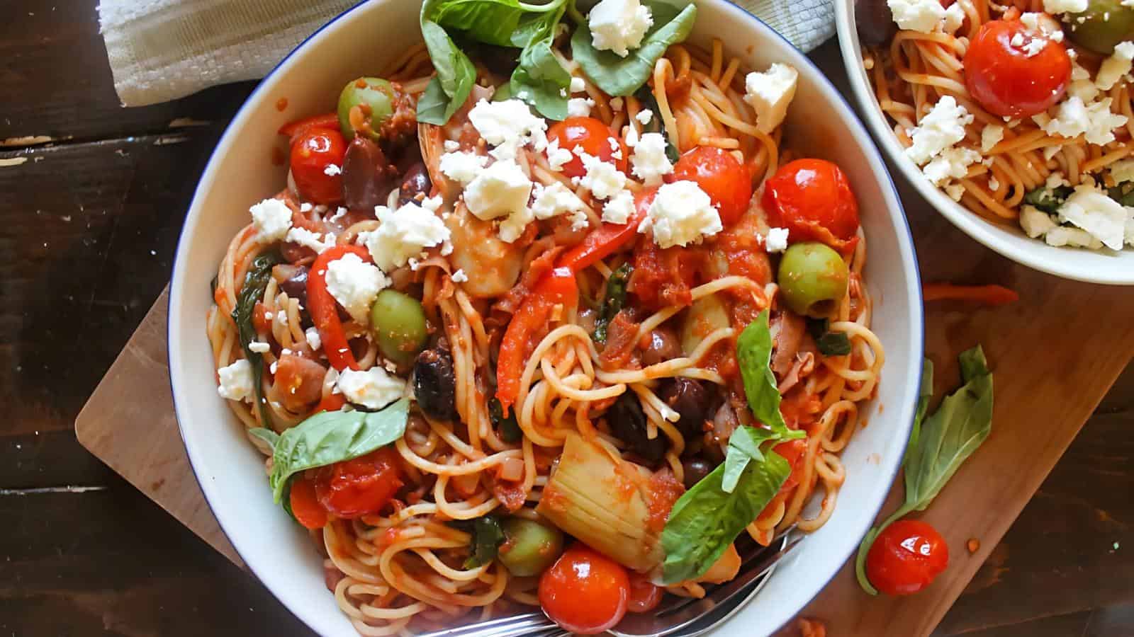 A bowl of spaghetti with cherry tomatoes, olives, spinach, basil leaves, artichokes, and crumbled feta cheese on a wooden board.