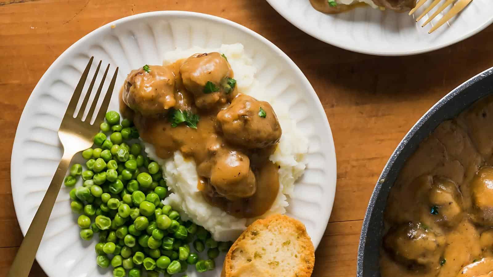 Plate with mashed potatoes, meatballs in brown sauce, and green peas, alongside a slice of bread and a gold fork, on a wooden table.