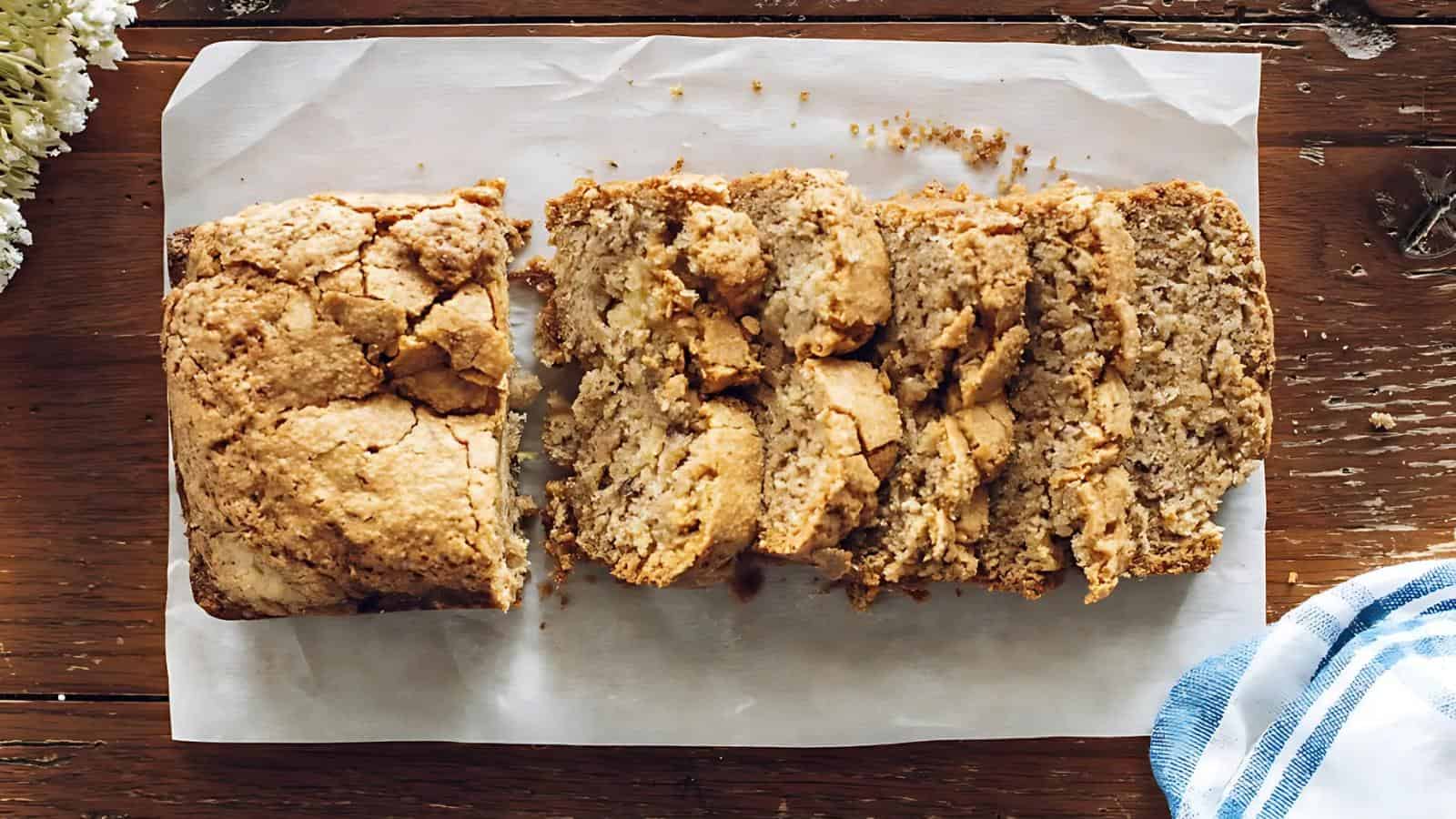 Sliced loaf of banana bread on parchment paper atop a wooden surface, with a blue and white cloth nearby.