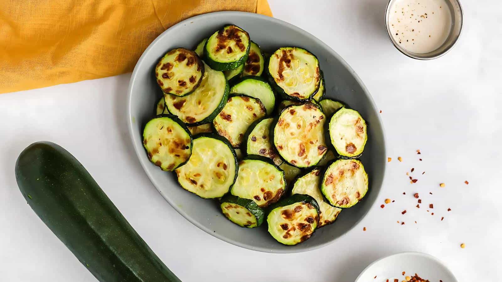 A plate of roasted zucchini slices on a white surface, surrounded by a whole zucchini, a yellow cloth, red pepper flakes, and a small cup of sauce.