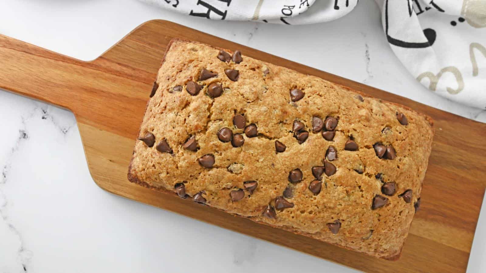A loaf of chocolate chip bread on a wooden cutting board, resting on a marble surface with a patterned cloth nearby.