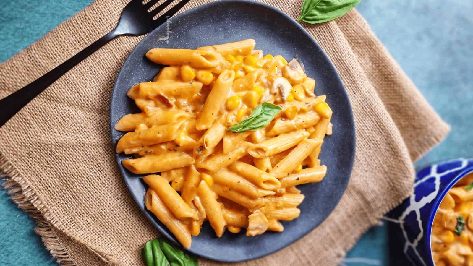 A plate of creamy penne pasta with corn and garnished with basil on a burlap mat. A fork and a bowl with similar pasta are nearby.