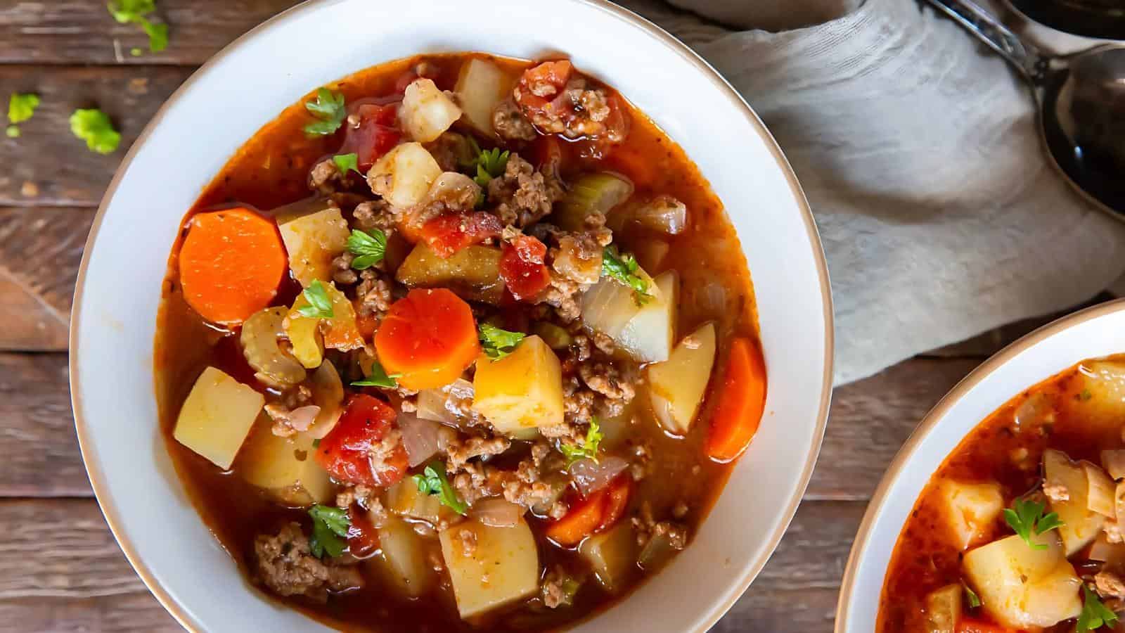 A bowl of hearty vegetable and beef stew with carrots, potatoes, tomatoes, and herbs, served on a wooden table next to a gray napkin and utensils.