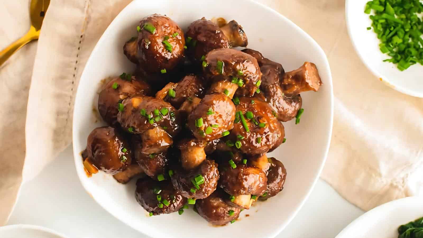 A white bowl filled with glazed mushrooms garnished with chopped green herbs.