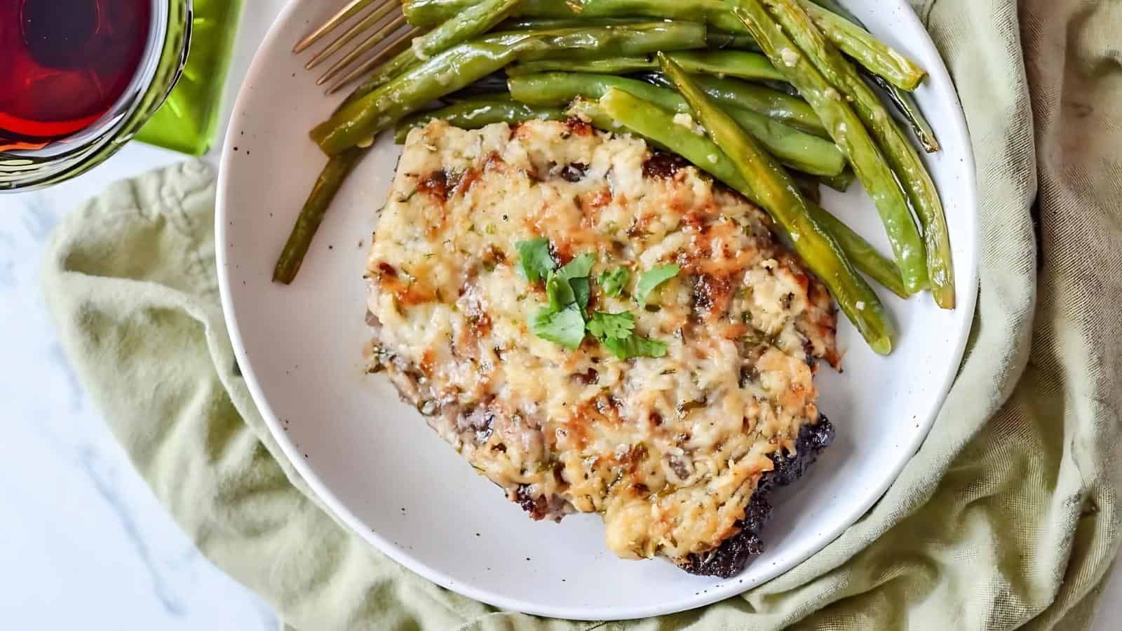 A plate with a slice of meatloaf topped with cheese and herbs, accompanied by green beans. A fork is next to the food. A glass with a red beverage is partially visible on the side.