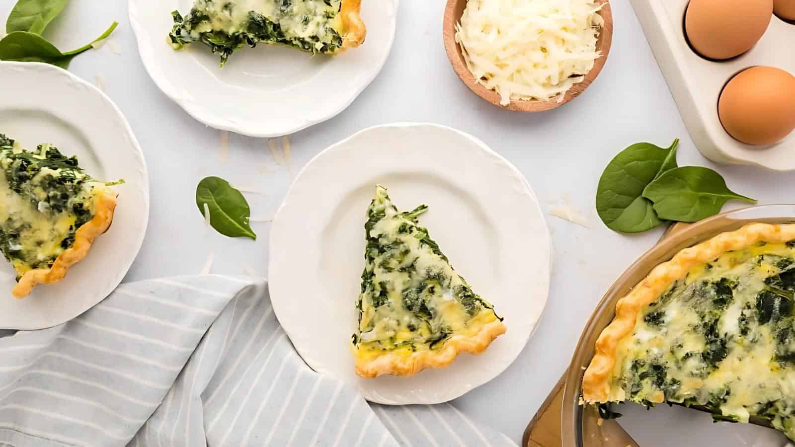 Slices of spinach and cheese quiche on white plates, surrounded by eggs, shredded cheese, and spinach leaves on a light surface.