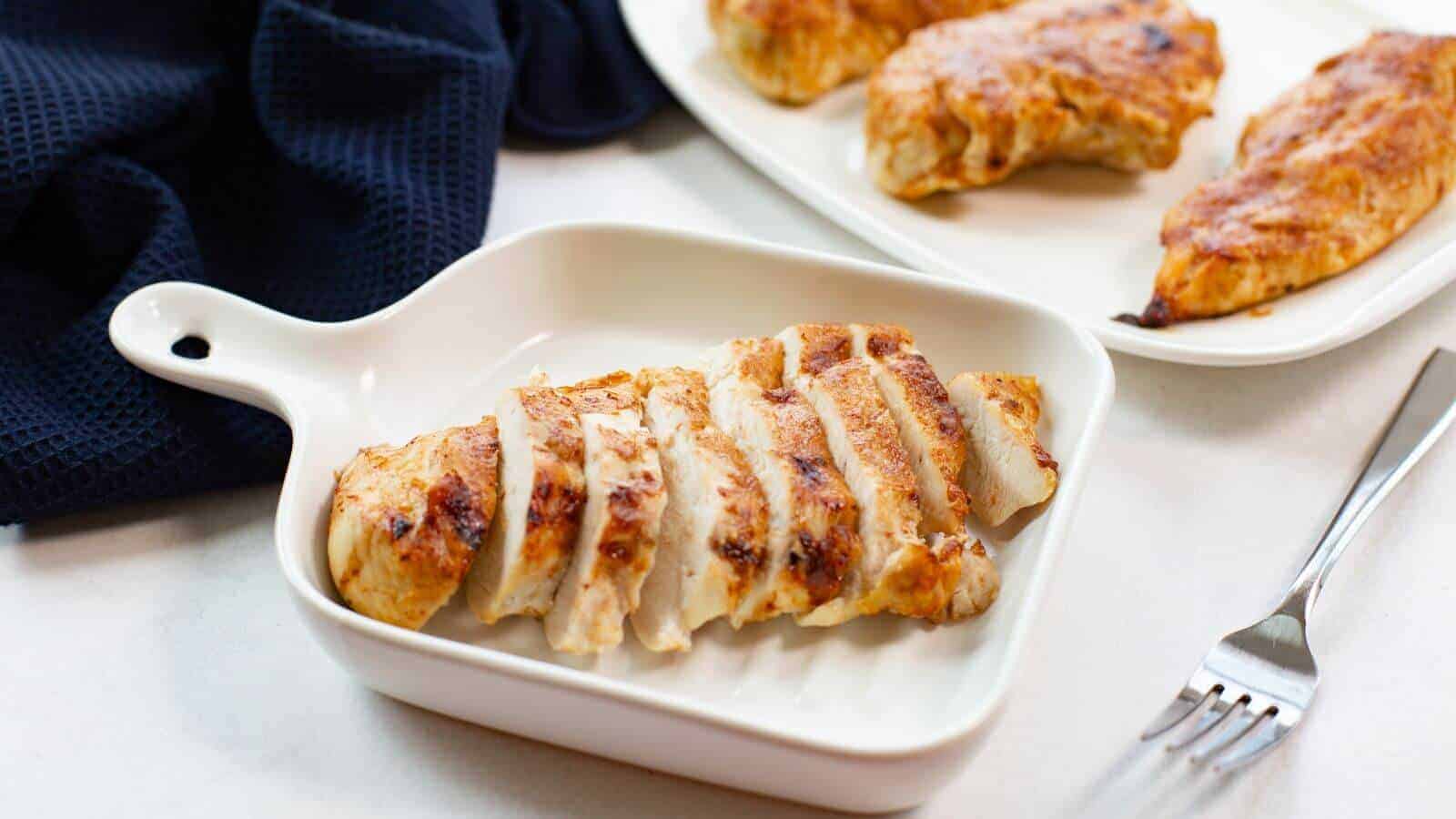 Sliced grilled chicken breast in a white dish, with whole chicken pieces on a separate plate in the background. A fork and a dark cloth are nearby on a white surface.