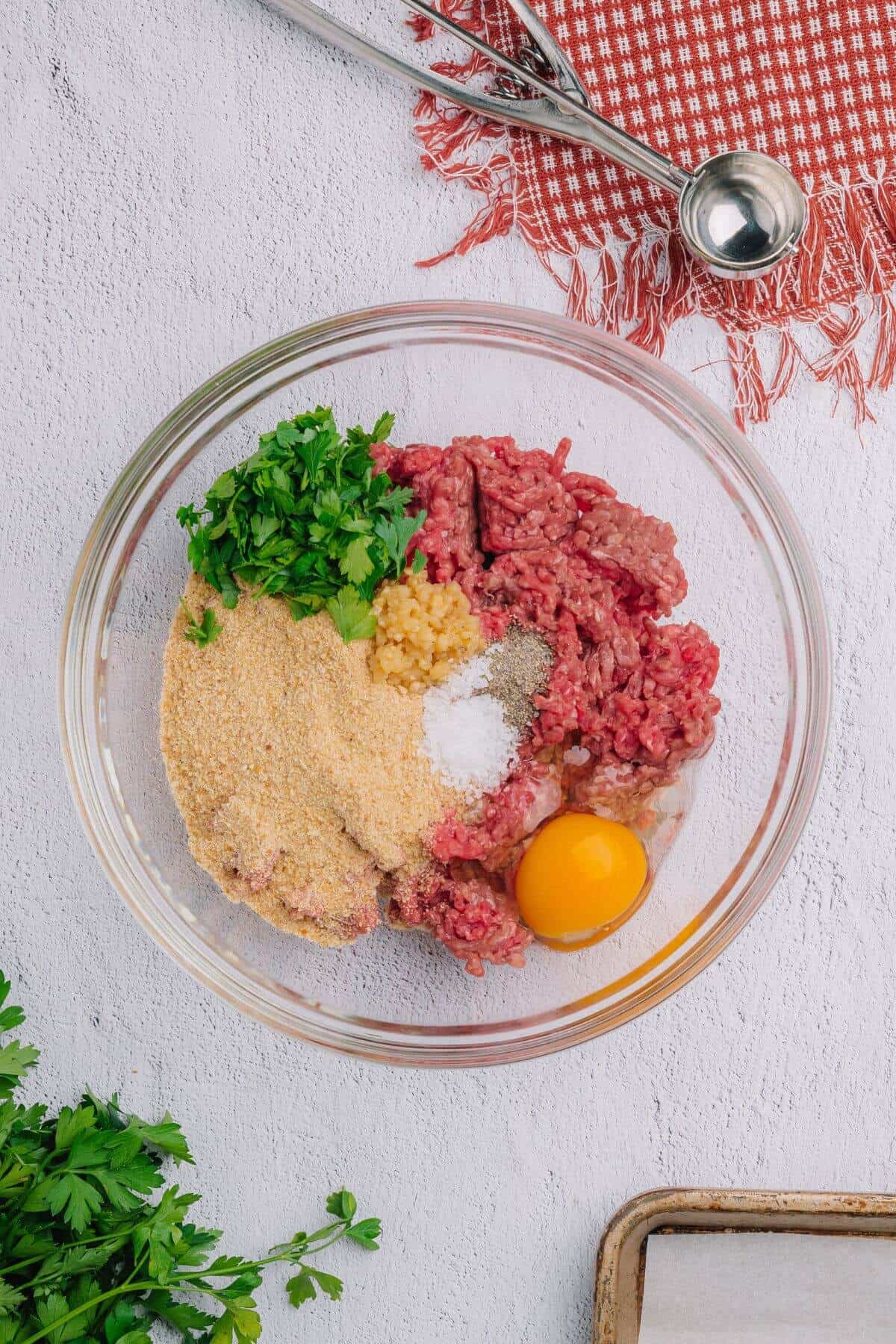 Glass bowl with ground meat, egg, breadcrumbs, parsley, salt, pepper, and minced garlic on a white surface with a red napkin and ice cream scoop nearby.