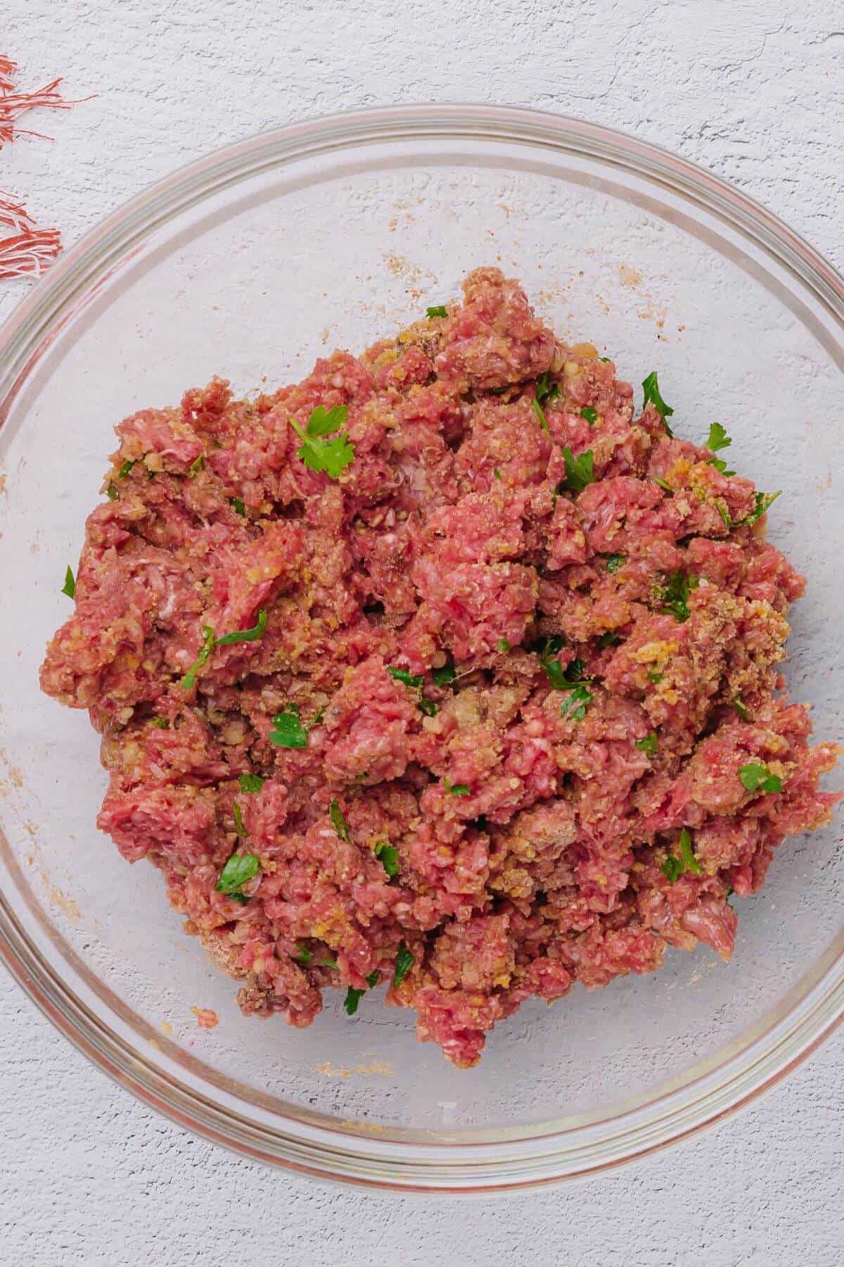 A clear glass bowl containing a mixture of raw ground beef and herbs on a white surface.