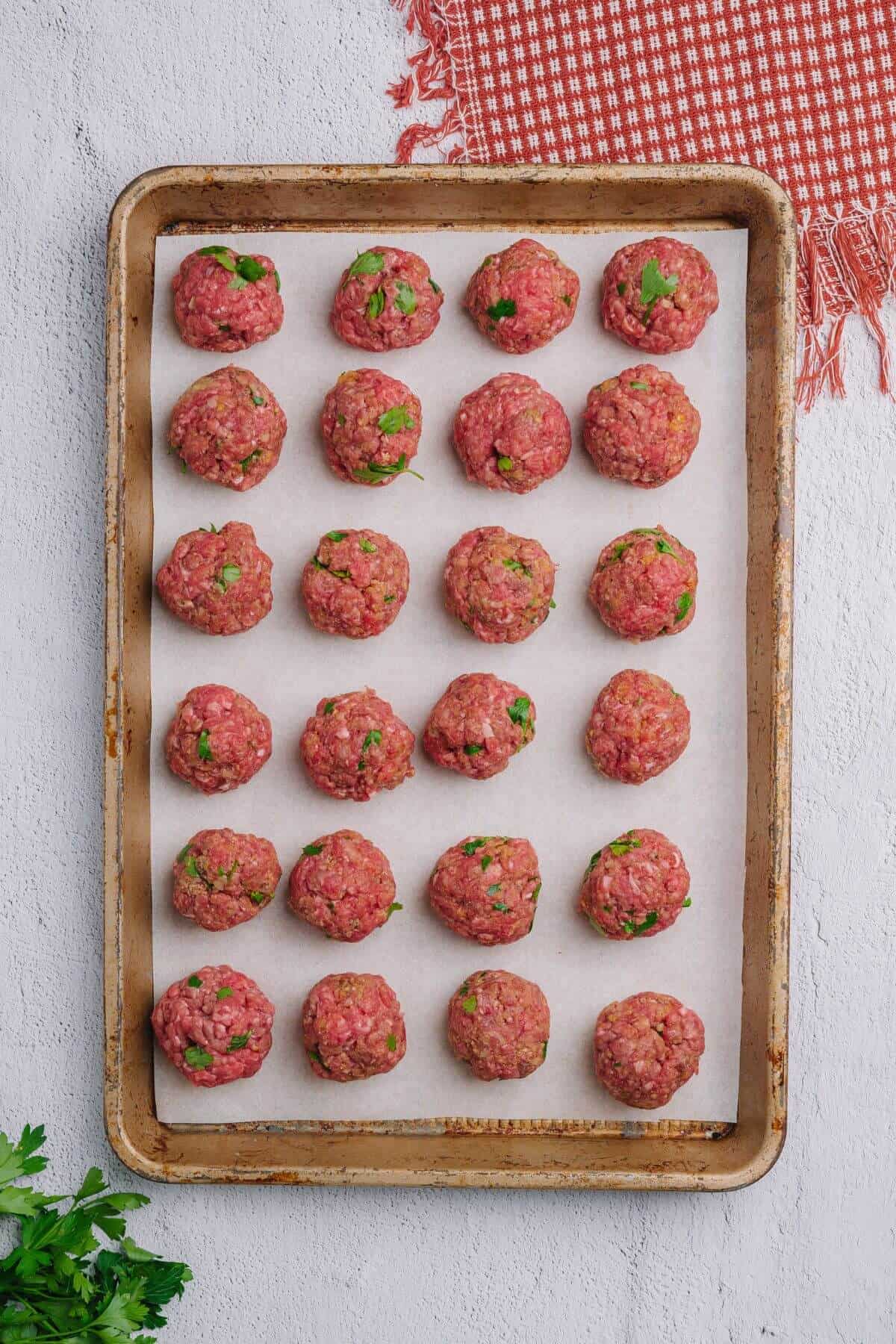 A baking sheet with evenly spaced raw meatballs on parchment paper, garnished with chopped herbs. There's a red and white cloth in the upper corner.