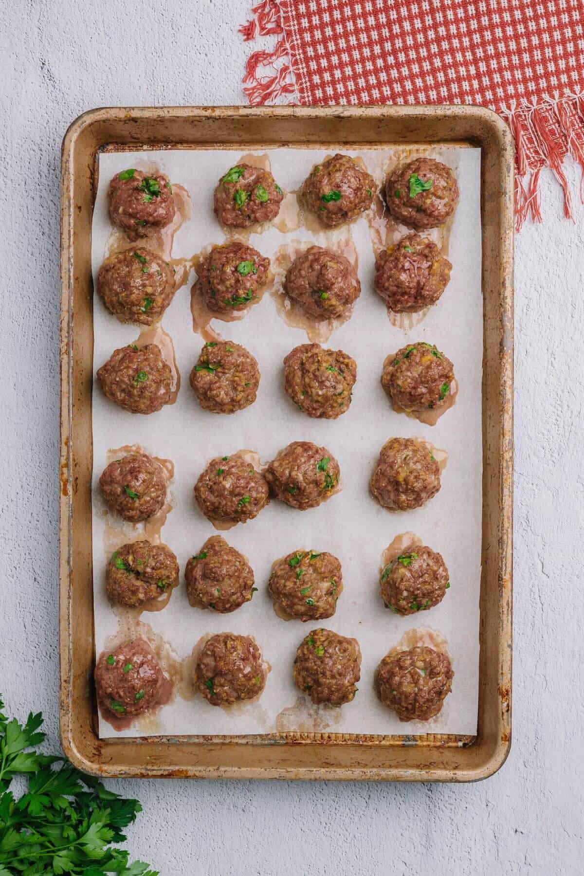 A baking tray with evenly spaced cooked meatballs on parchment paper, garnished with chopped herbs. A red-checkered cloth is partially visible in the corner.