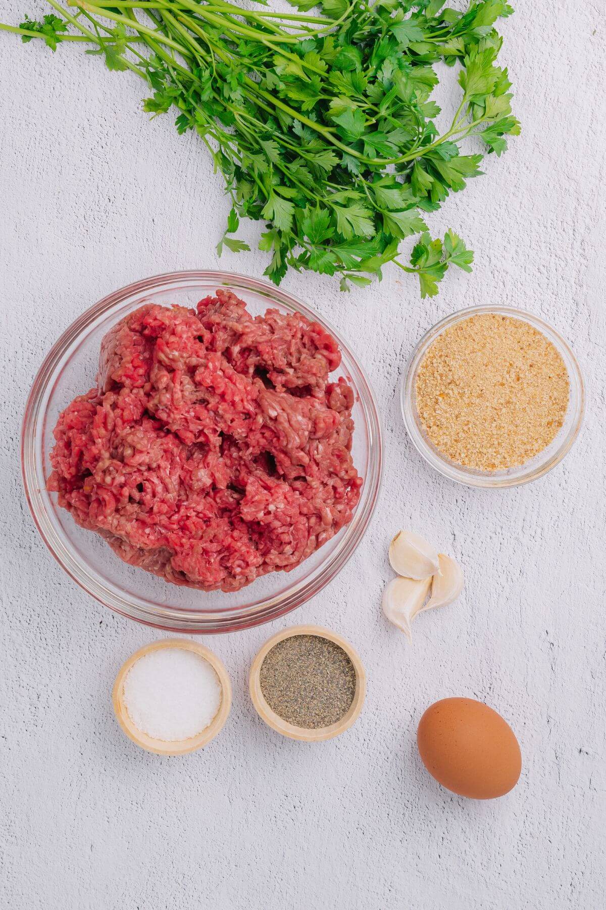 Ground meat in a bowl, with garlic cloves, an egg, bread crumbs, salt, pepper, and parsley arranged around it on a white surface.