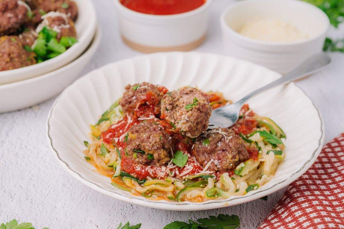 A plate of spaghetti with meatballs and tomato sauce, garnished with parsley and grated cheese. A fork is placed in the dish. Two bowls in the background contain sauce and more cheese.