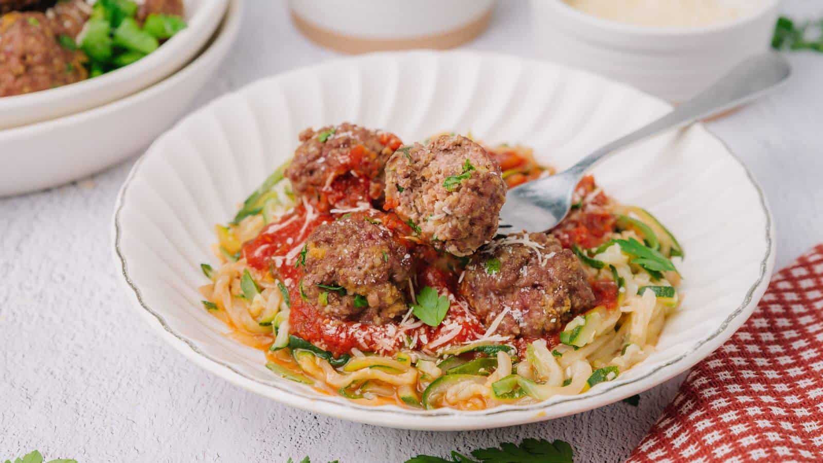 A plate of zucchini noodles topped with tomato sauce, meatballs, and grated cheese, garnished with parsley. A fork rests on the plate.