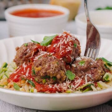 A plate of zucchini noodles topped with meatballs, marinara sauce, and grated cheese, garnished with parsley. A fork is piercing a meatball.