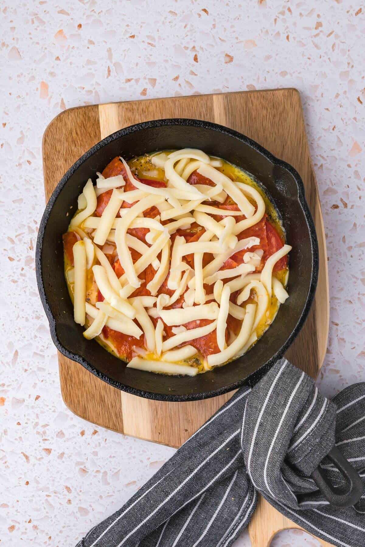 A cast iron skillet on a wooden board contains sliced tomatoes topped with shredded cheese. A striped cloth is nearby.