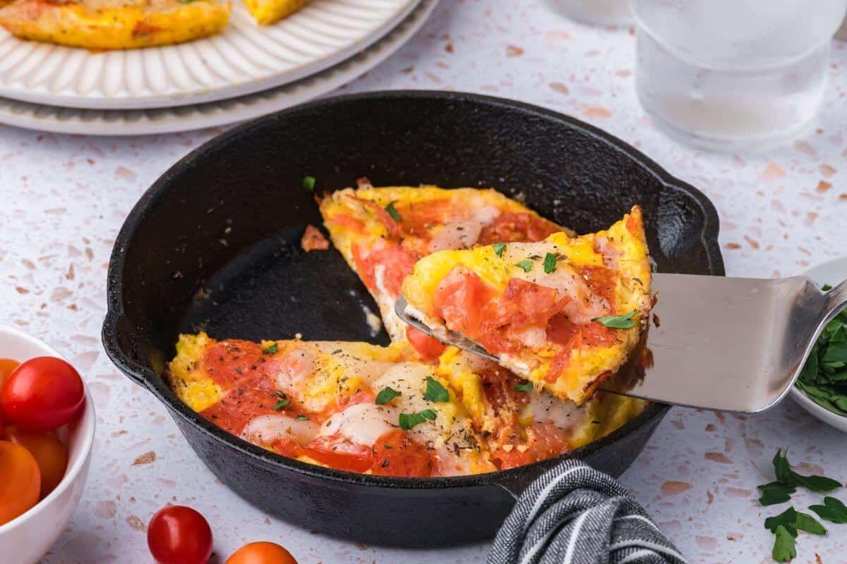 A slice of tomato and cheese frittata is being lifted from a cast iron pan. The table also holds a bowl of cherry tomatoes and a plate with a napkin.
