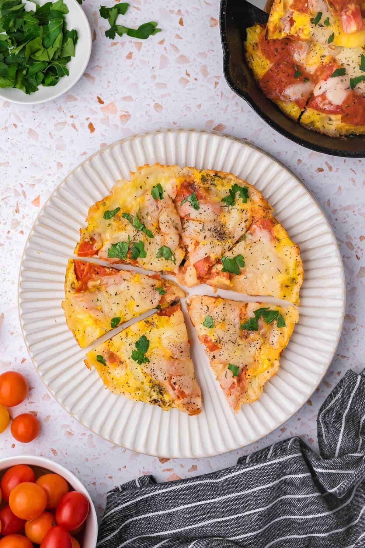 A sliced omelette topped with tomatoes and herbs on a white plate. Nearby are a bowl of cherry tomatoes, a dish of herbs, and another omelette in a skillet. A striped cloth is also visible.