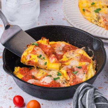 A slice of pepperoni pizza is being lifted from a skillet. The pizza features cheese and herbs, with a background of a glass of water and a small plate. Cherry tomatoes are nearby.
