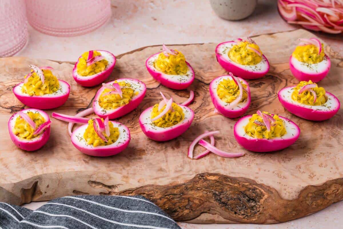 Deviled eggs with bright pink edges topped with yellow filling and garnished with sliced red onions, displayed on a rustic wooden board.