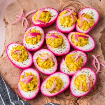 Deviled eggs with bright pink edges, yellow yolk filling, and garnished with dill and pickled onion, arranged on a wooden surface.