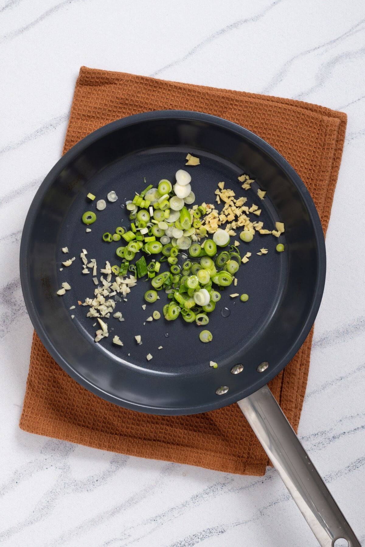 A frying pan with chopped garlic and green onions on a brown cloth, placed on a marble surface.