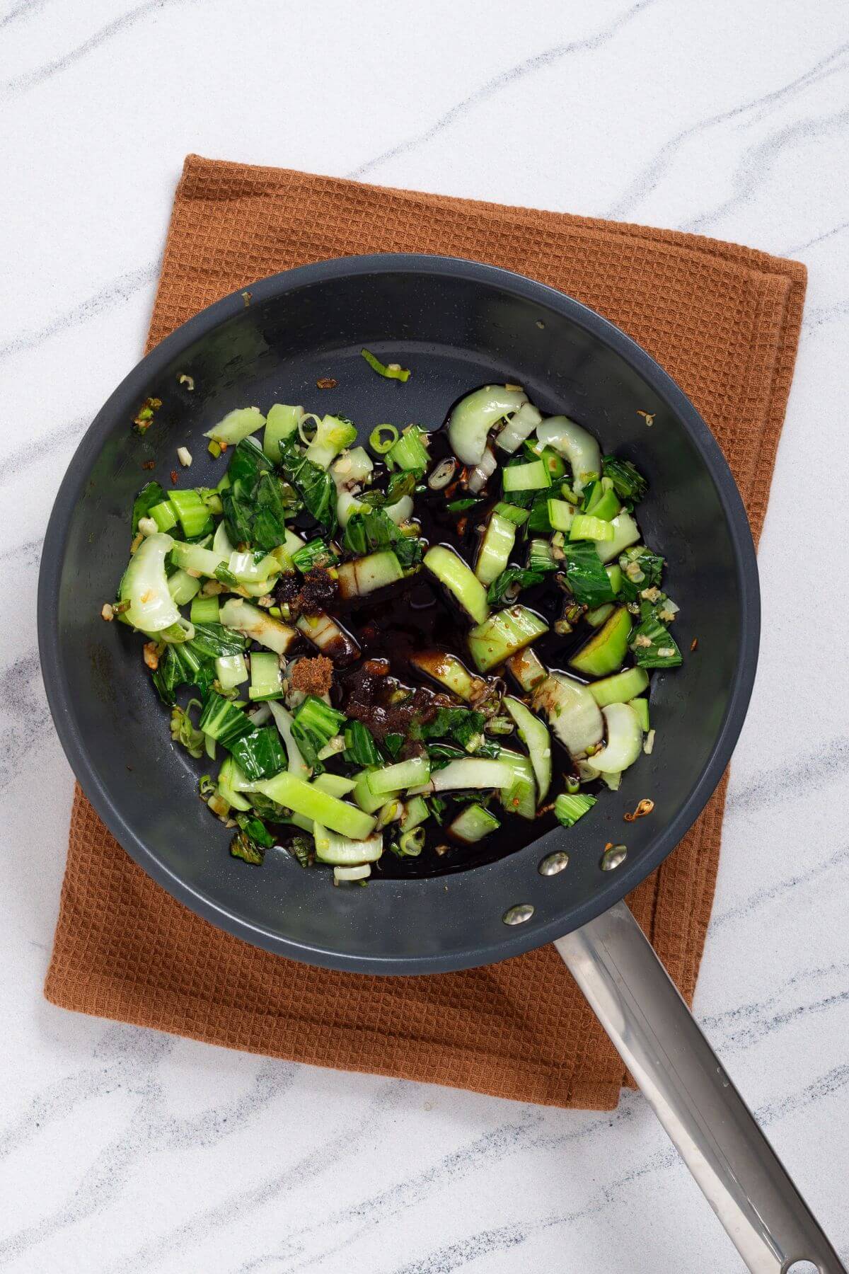 A frying pan on a brown mat contains chopped green vegetables and soy sauce on a white marble surface.