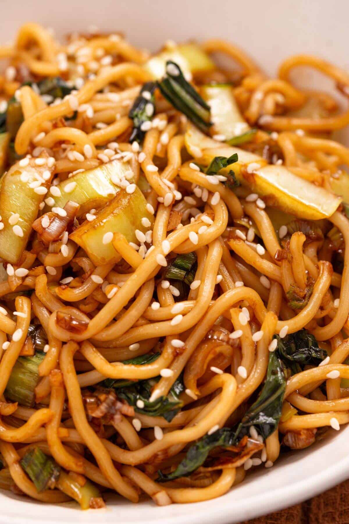 Close-up of a plate of sesame noodles with greens and sliced vegetables, topped with sesame seeds.