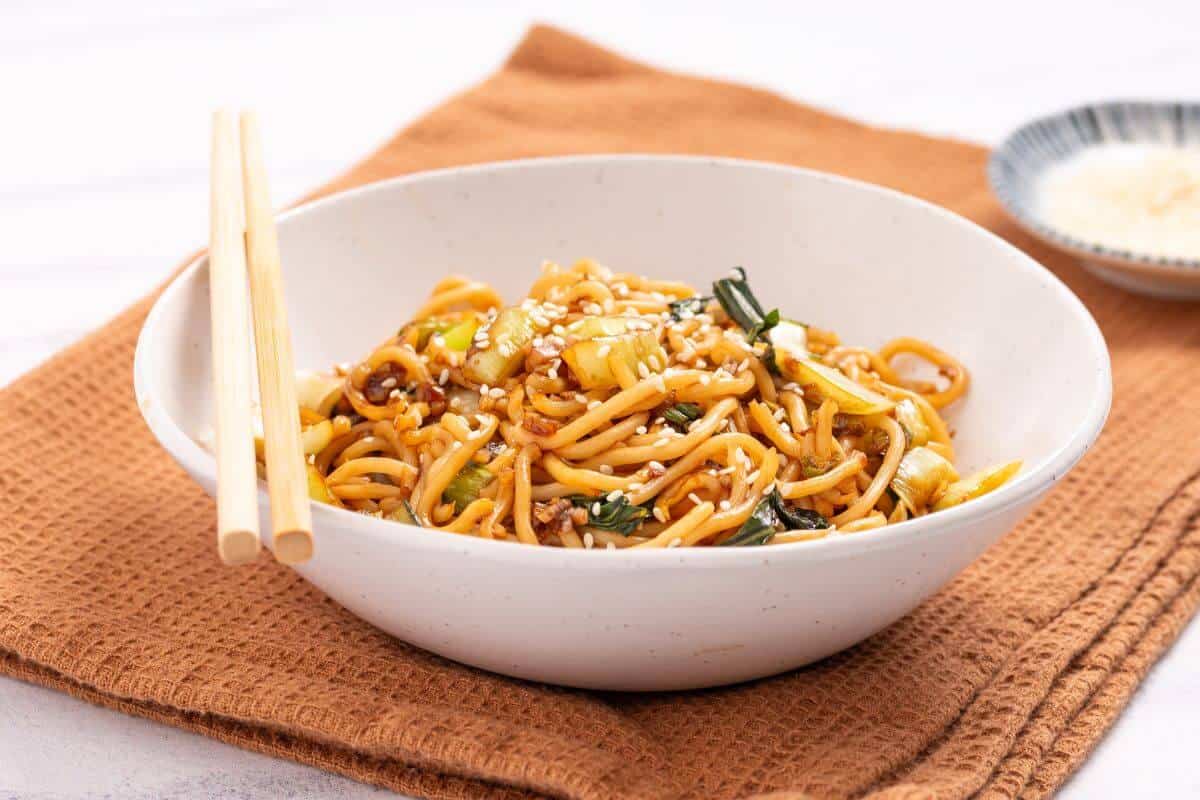 Bowl of stir-fried noodles with vegetables and sesame seeds, placed on a brown cloth with wooden chopsticks resting on the bowl's edge.