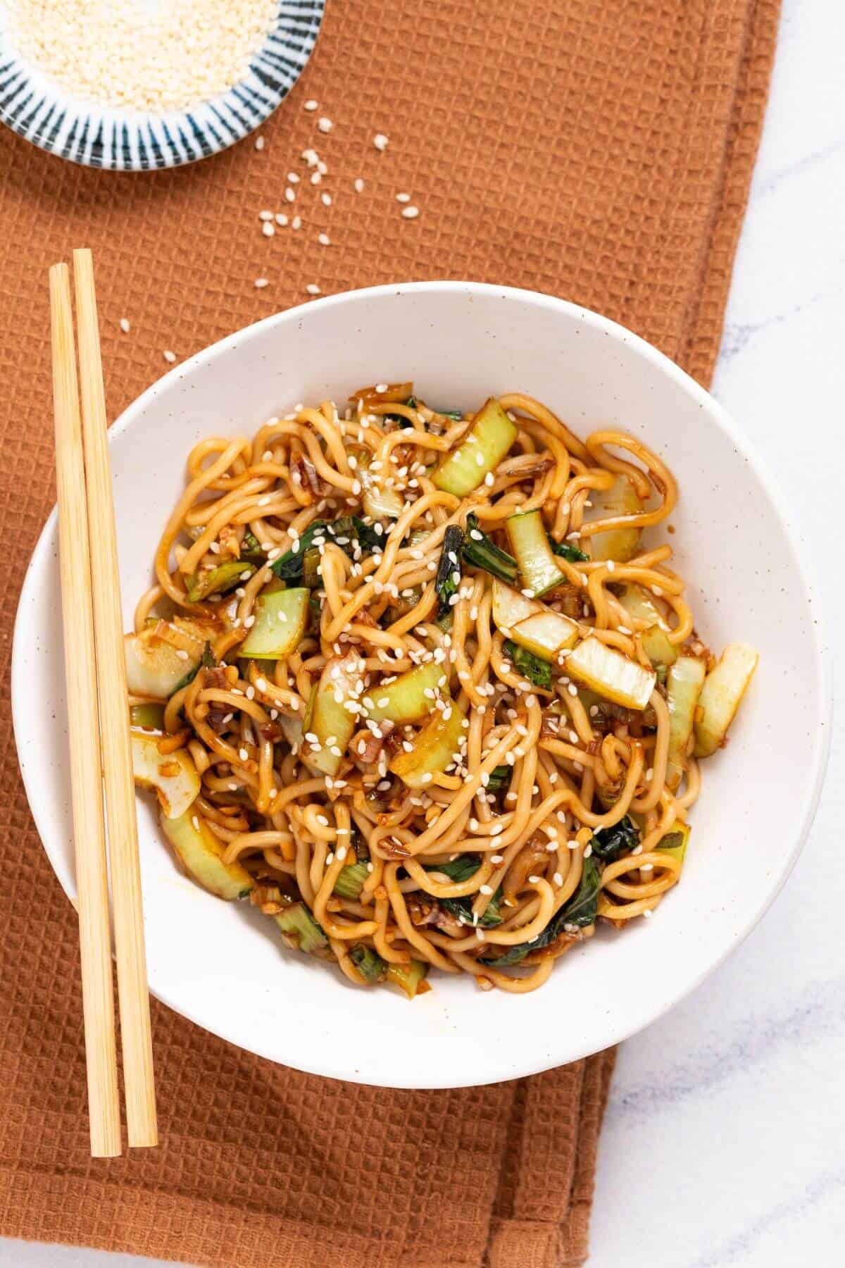 A bowl of stir-fried noodles with vegetables, topped with sesame seeds. Chopsticks rest on the bowl. A small dish of sesame seeds is in the background on a brown cloth.