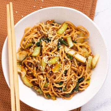 A bowl of stir-fried noodles with vegetables, topped with sesame seeds, and served with chopsticks on a brown textured napkin.