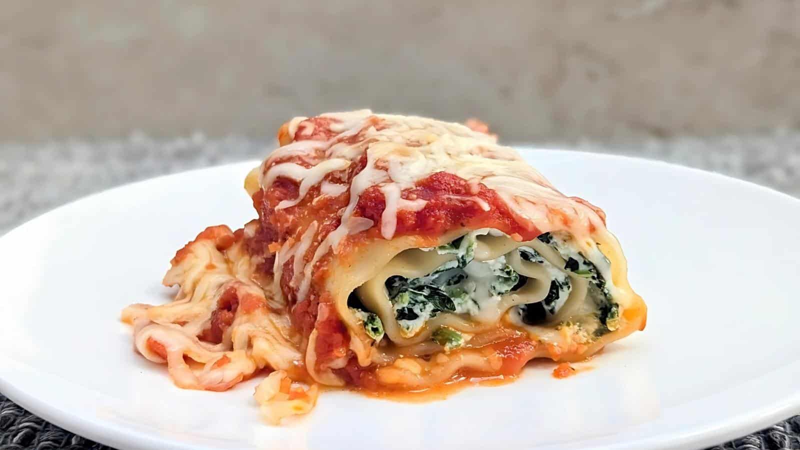 A piece of rolled lasagna with spinach and cheese filling on a white plate, topped with melted cheese and tomato sauce.