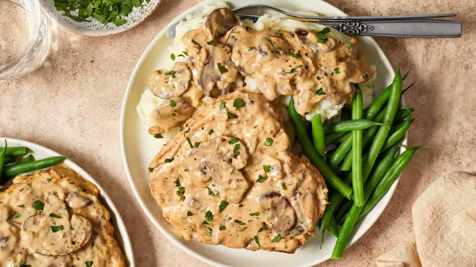 Plate of pork chops with creamy mushroom sauce, mashed potatoes, and green beans, garnished with chopped parsley. A fork and knife are placed on the plate.