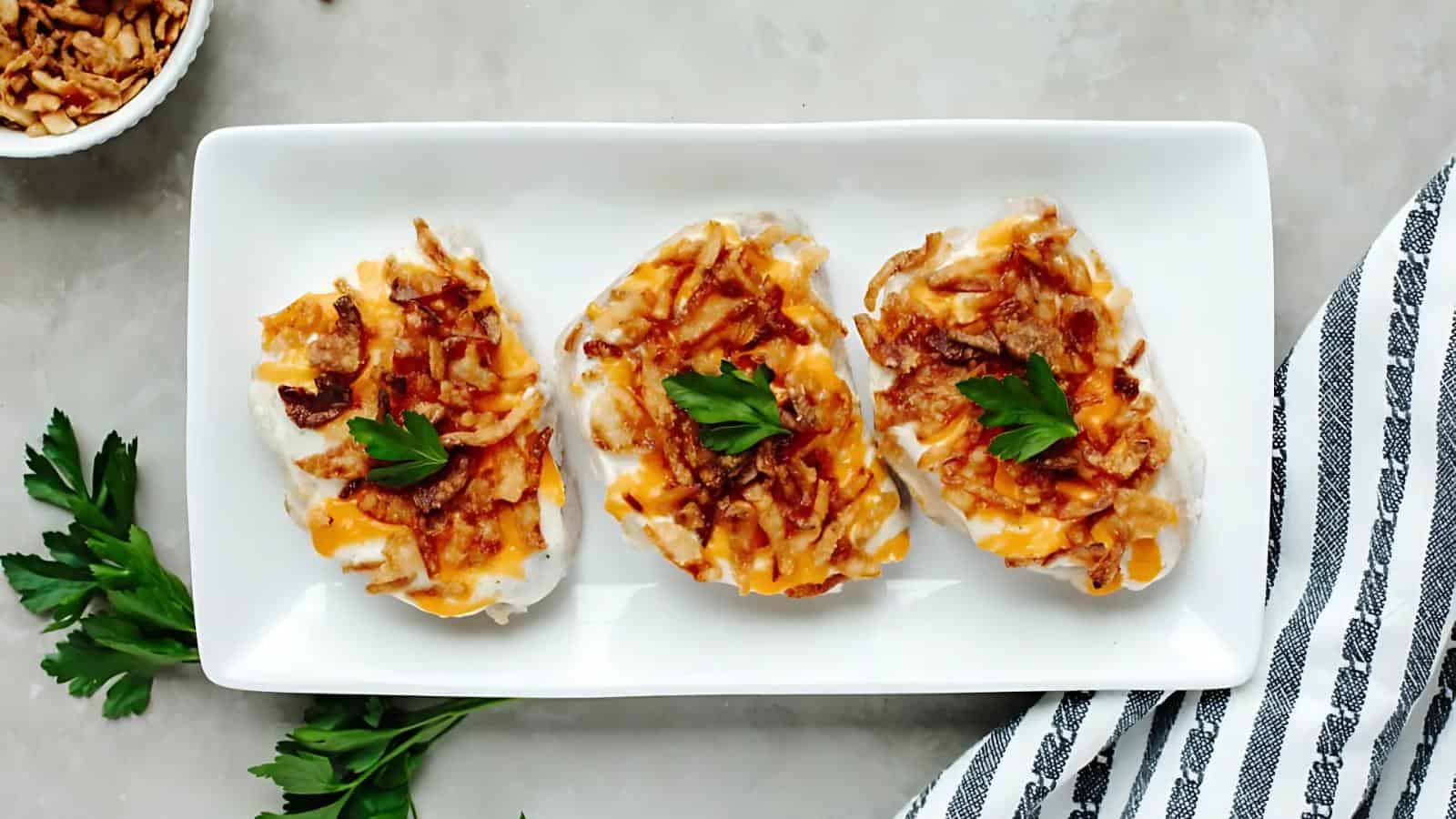 Three pieces of cheesy baked chicken breast topped with crispy onions and garnished with parsley on a rectangular white plate.