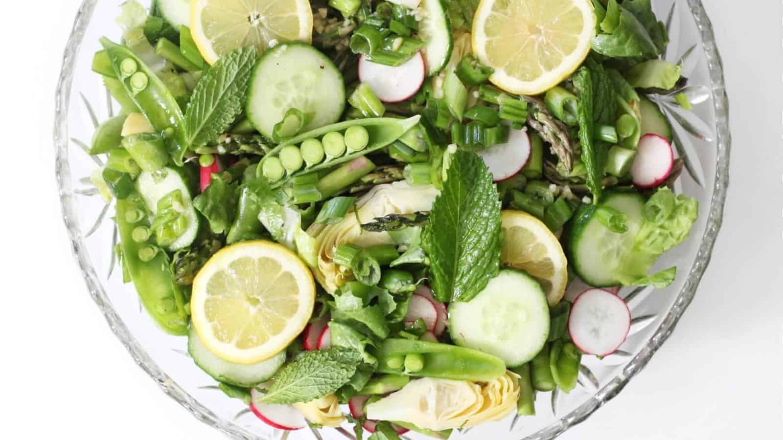 A glass bowl filled with a salad containing lemon slices, cucumber, snap peas, radishes, artichoke hearts, asparagus, green onions, lettuce, and mint leaves.