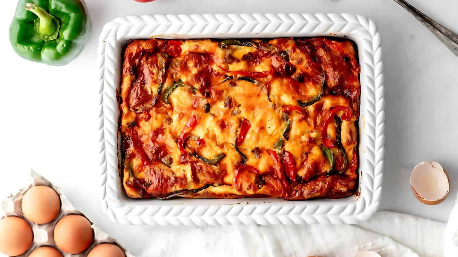 A baked casserole in a white dish with visible cheese and vegetables. Surrounding items include eggs, egg shells, a green bell pepper, and a white cloth on a marble surface.