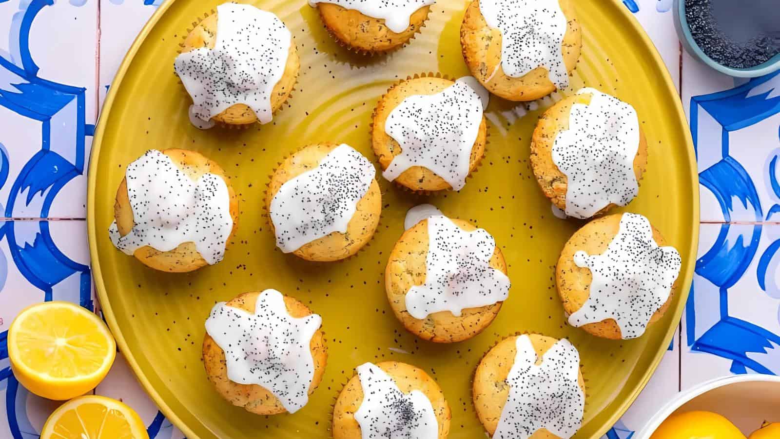 A yellow plate with twelve cupcakes topped with white icing and sprinkled with poppy seeds, surrounded by lemon halves and a bowl of poppy seeds on a patterned surface.