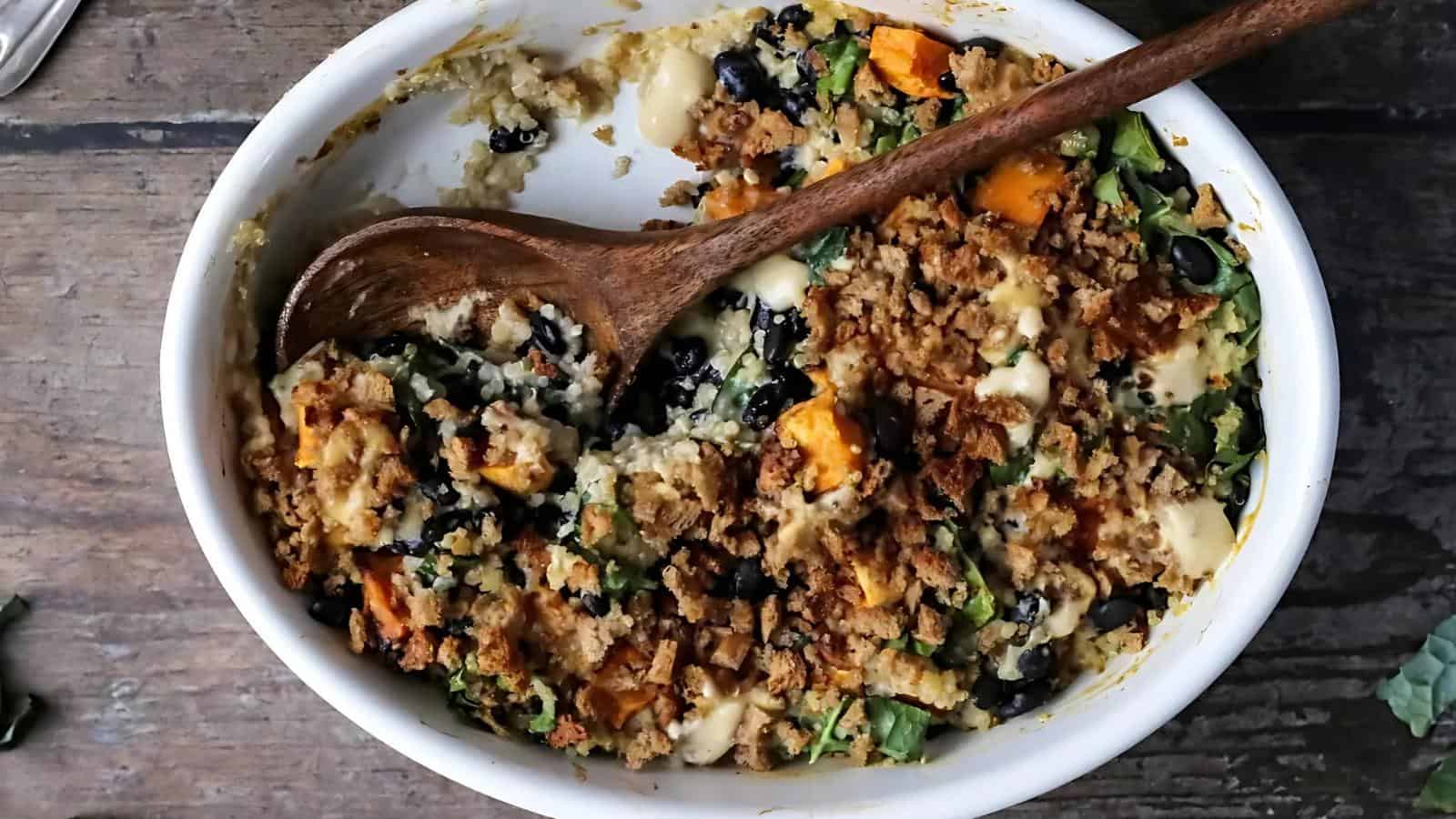 A baked casserole dish containing a mixture of vegetables, beans, leafy greens, and a breadcrumb topping, with a wooden spoon resting inside.