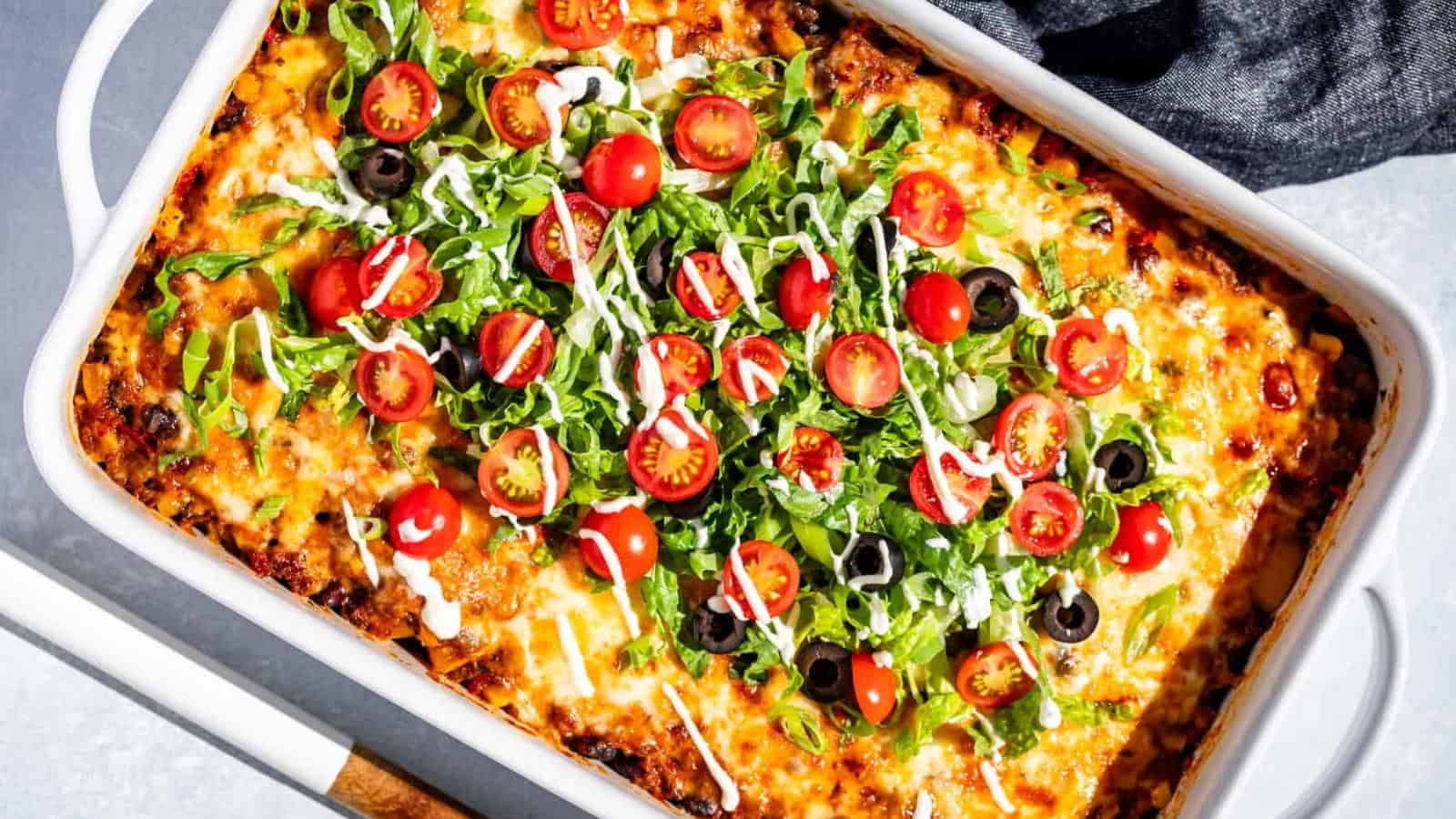 A baked casserole topped with melted cheese, shredded lettuce, sliced cherry tomatoes, black olives, and drizzled with a white sauce in a white rectangular dish.