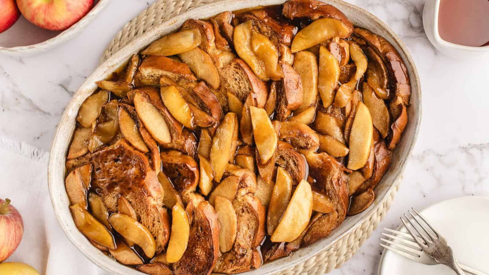 A baked dish of sliced apples layered on brown bread pudding in an oval dish on a marble surface, with whole apples and a fork nearby.