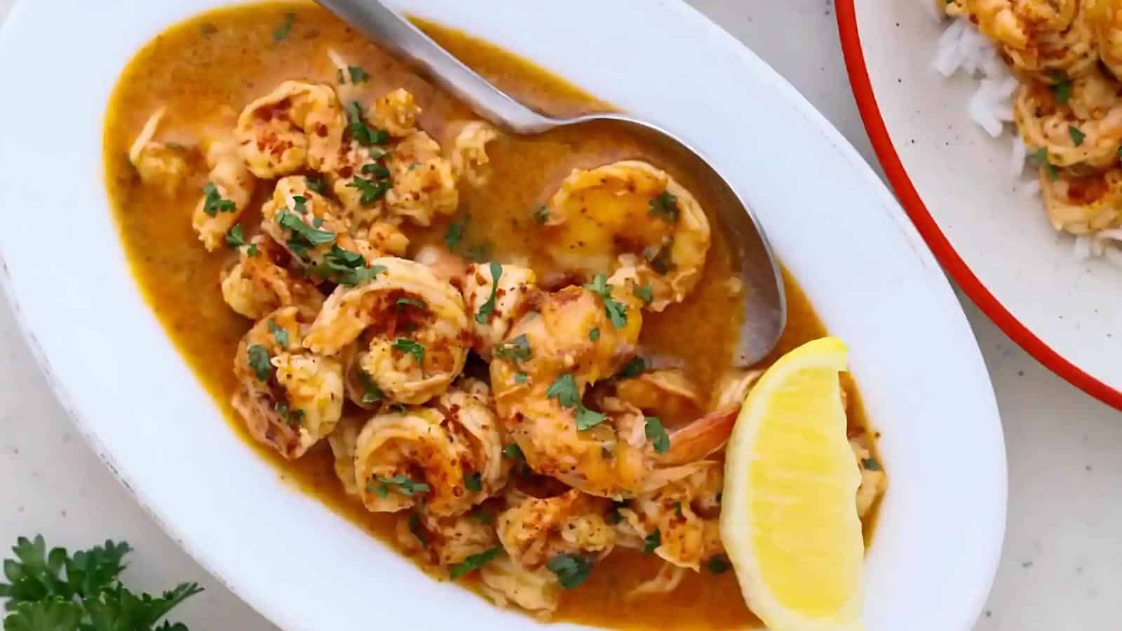 A dish of cooked shrimp in a spicy, orange sauce is garnished with chopped herbs and a lemon wedge. A spoon is placed on the side.