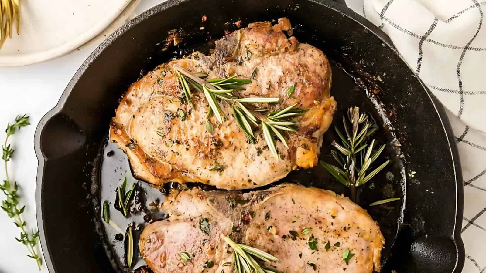 Two cooked pork chops garnished with fresh rosemary and herbs in a black cast iron skillet.