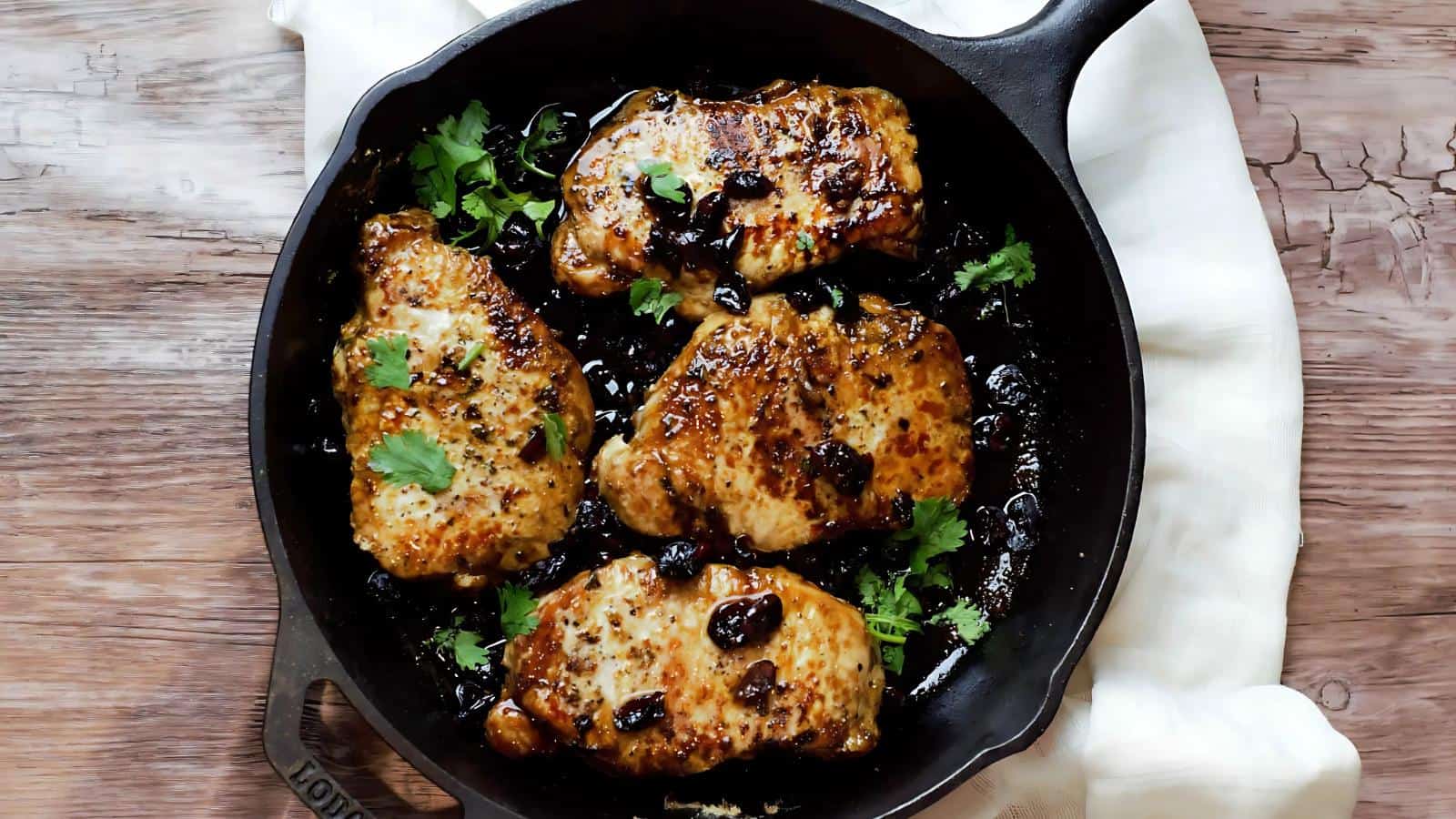 Four cooked chicken thighs with herbs and olives are arranged in a black cast iron skillet on a wooden surface.