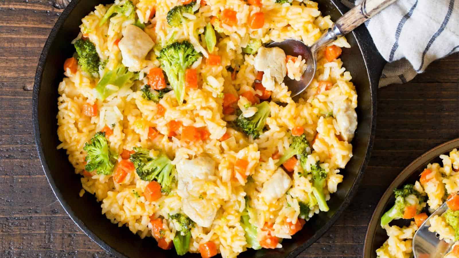 A bowl of rice mixed with broccoli, carrots, and chicken pieces, with a spoon resting in the dish on a wooden surface.