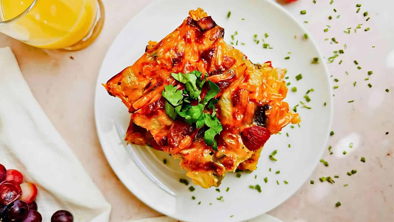 Plate of baked pasta topped with herbs, next to a glass of orange juice and grapes on a light surface.