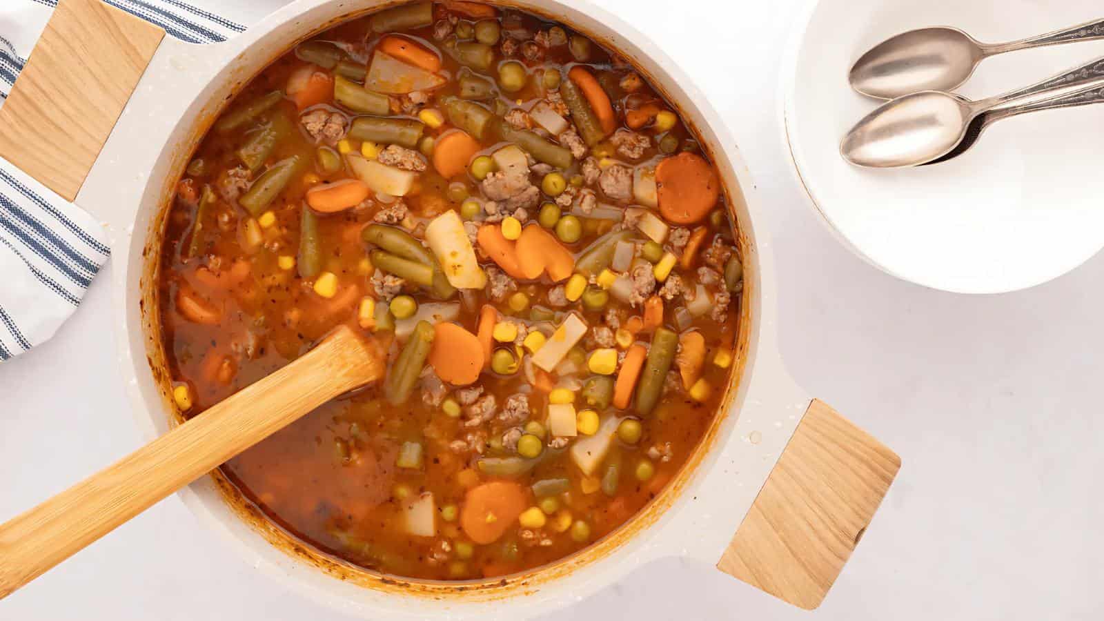 A pot of vegetable soup with carrots, green beans, corn, peas, and ground meat, stirred with a wooden spoon, next to an empty bowl and two spoons on a white surface.