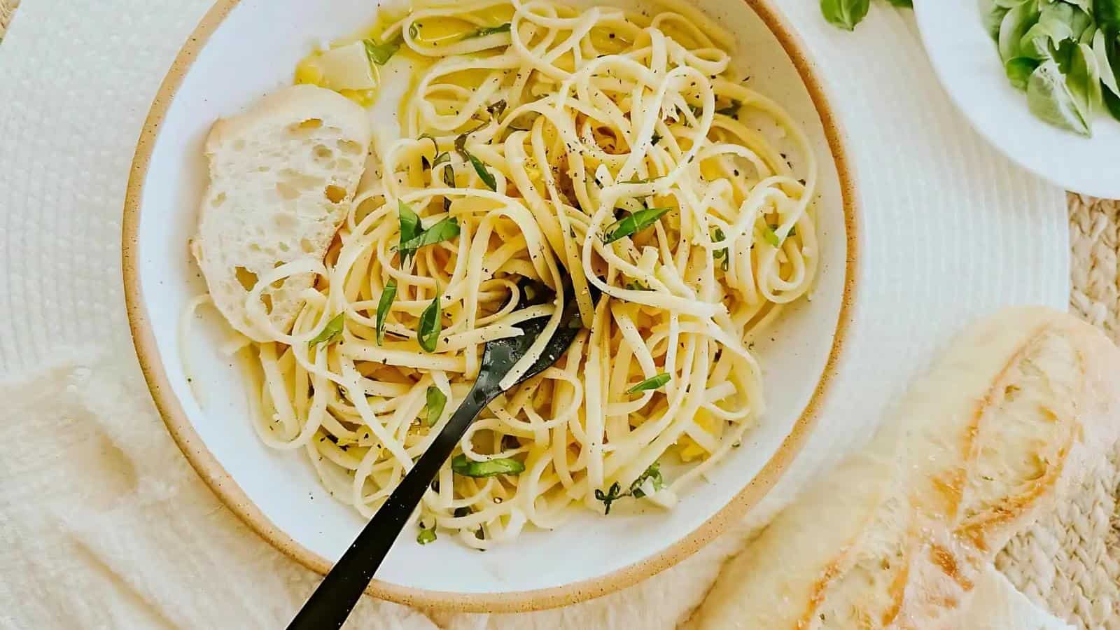 A bowl of spaghetti with herbs and broth, topped with a slice of bread, with a fork, a baguette, and a plate of greens on the side.