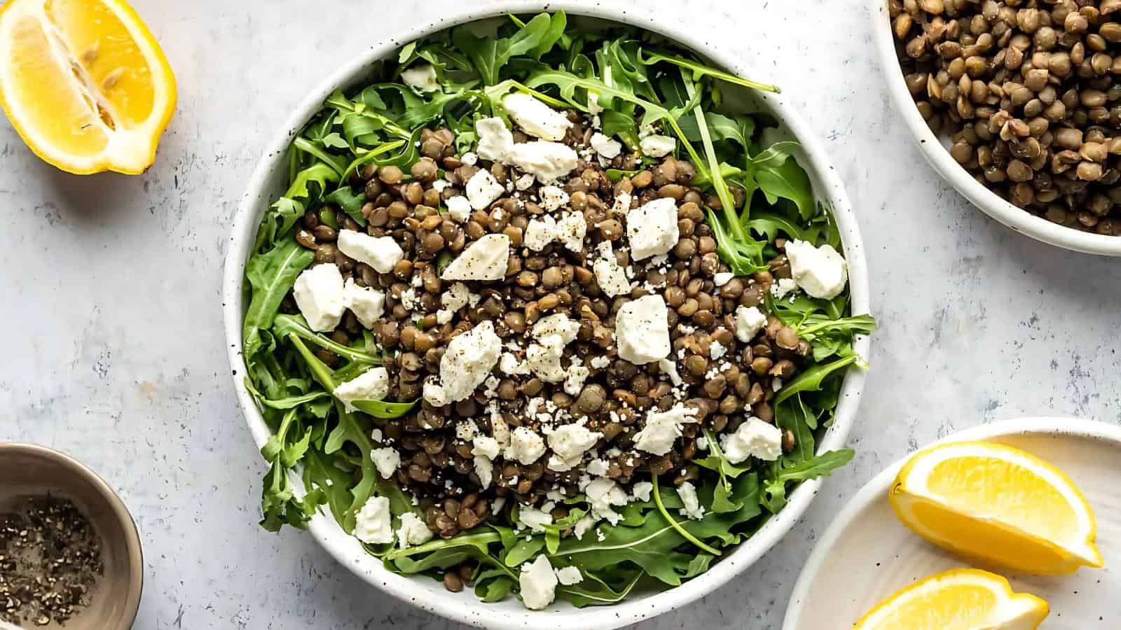 A bowl of arugula topped with cooked lentils and crumbled feta cheese, surrounded by lemon wedges and a bowl of lentils.