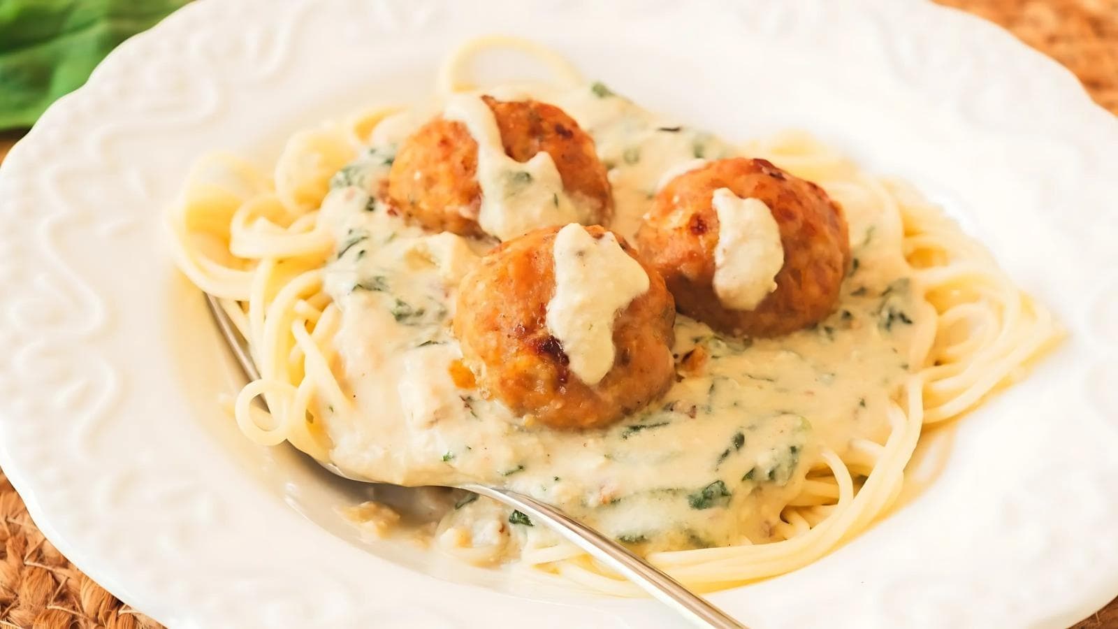 A plate of spaghetti topped with three meatballs and creamy white sauce, with a fork resting on the side.